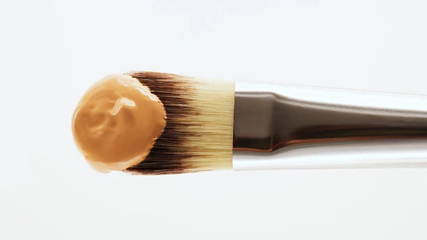Close-up of a flat foundation makeup brush with a dollop of liquid foundation on top against a white background.