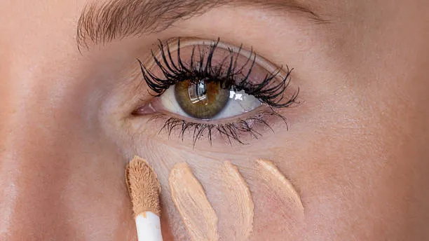Close-up of a woman applying liquid concealer in dots/stripes under her eye using a wand applicator.