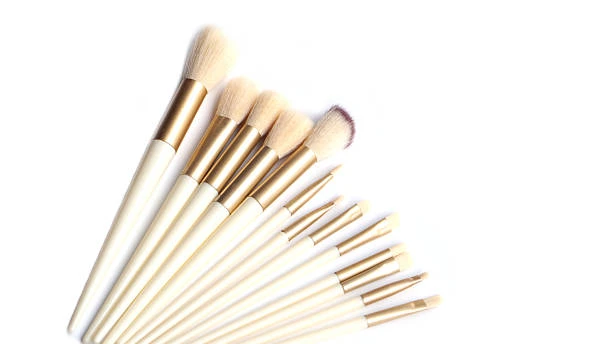 Set of 12 professional makeup brushes with cream-colored handles and gold ferrules arranged on a white background.