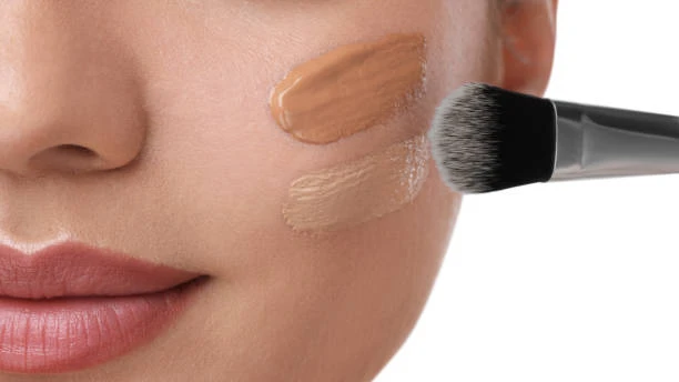 Close-up of a woman's face with two foundation swatches on her cheek, using a makeup brush to apply.